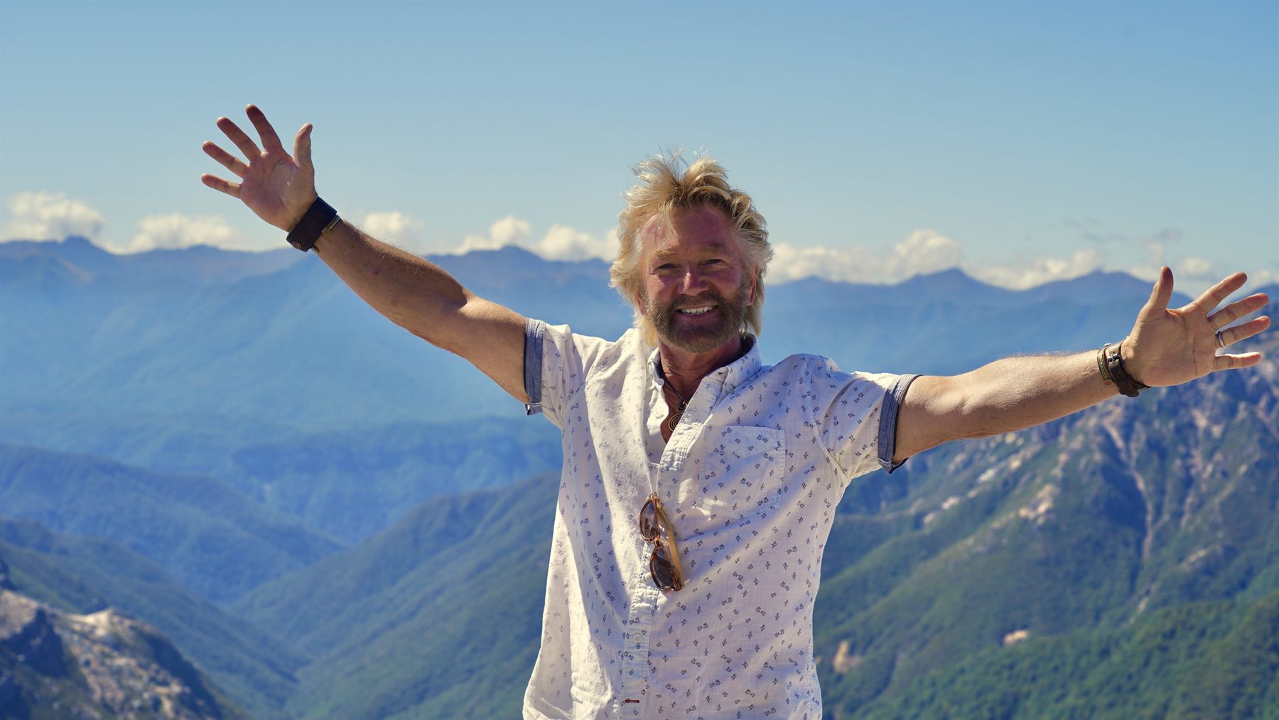 Noel Edmonds on a mountaintop with his arms outstretched, blue sky and hills behind him